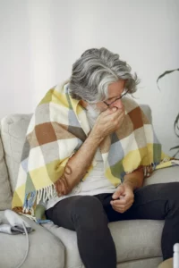 Elderly man with silver hair sitting on a couch, possibly with whooping cough holding his forehead in discomfort.